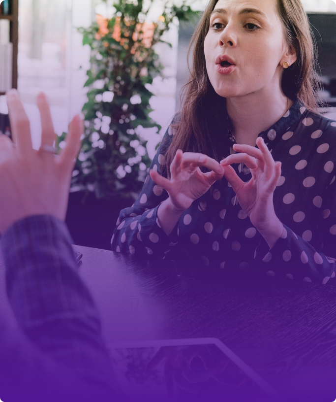 Only the hands of one of the 2 people speaking in sign language and sitting opposite each other are visible. In front of the person whose hands are visible, there is a woman with brown long hair, brown eyes, wearing a black and white rounded shirt and making the letter ‘B’ with her hands